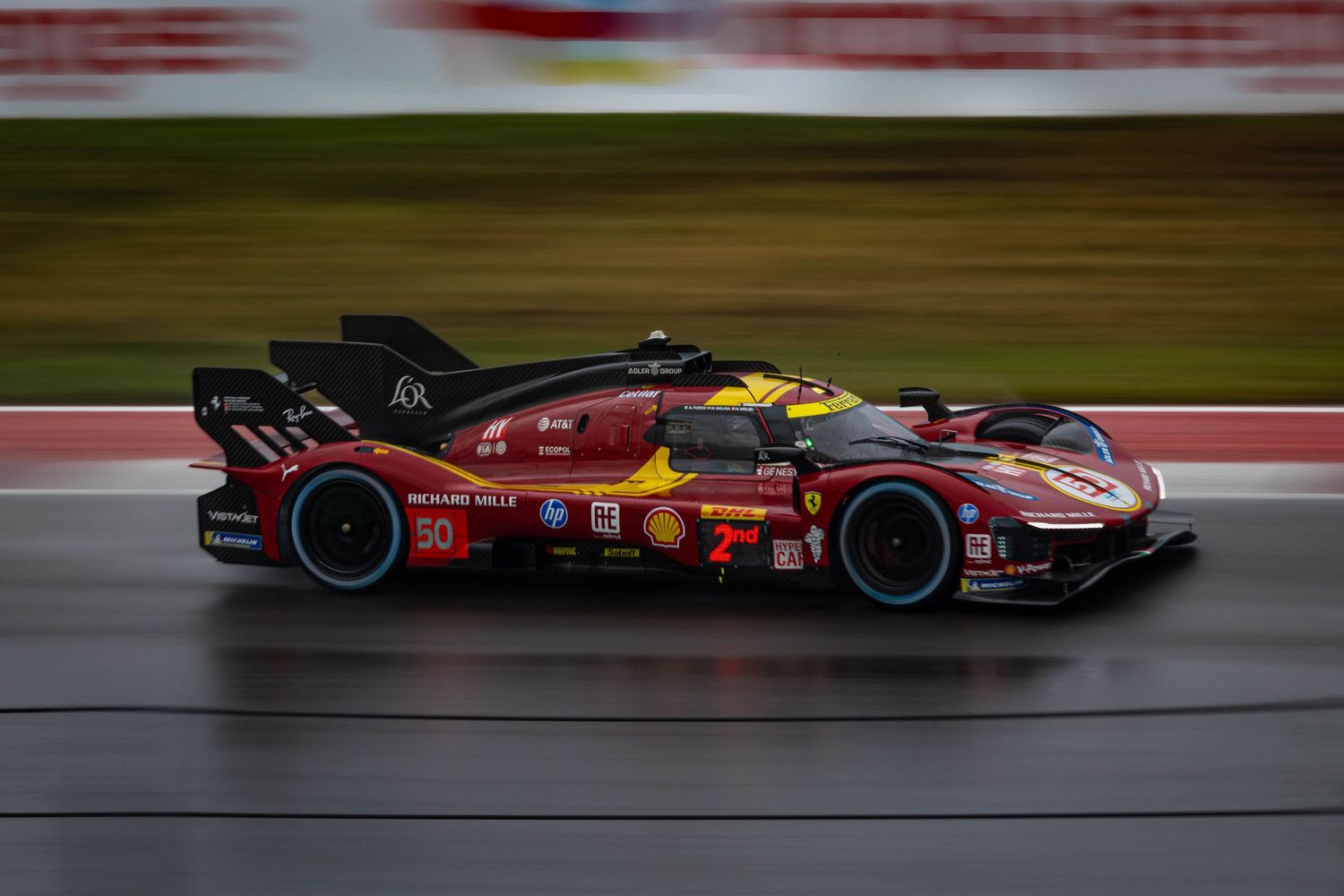 Ferrari 499P LMH prototype race car with Shell livery races on wet track with motion blur.