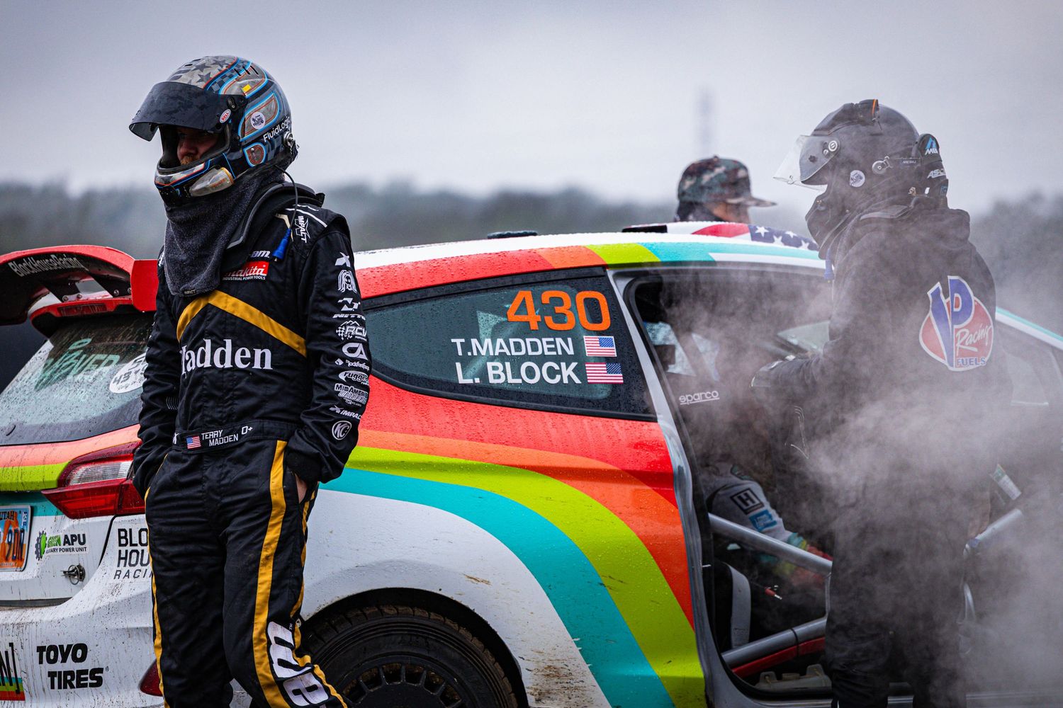 Rally driver T. Madden standing beside a rainbow-liveried race car surrounded by tire smoke.