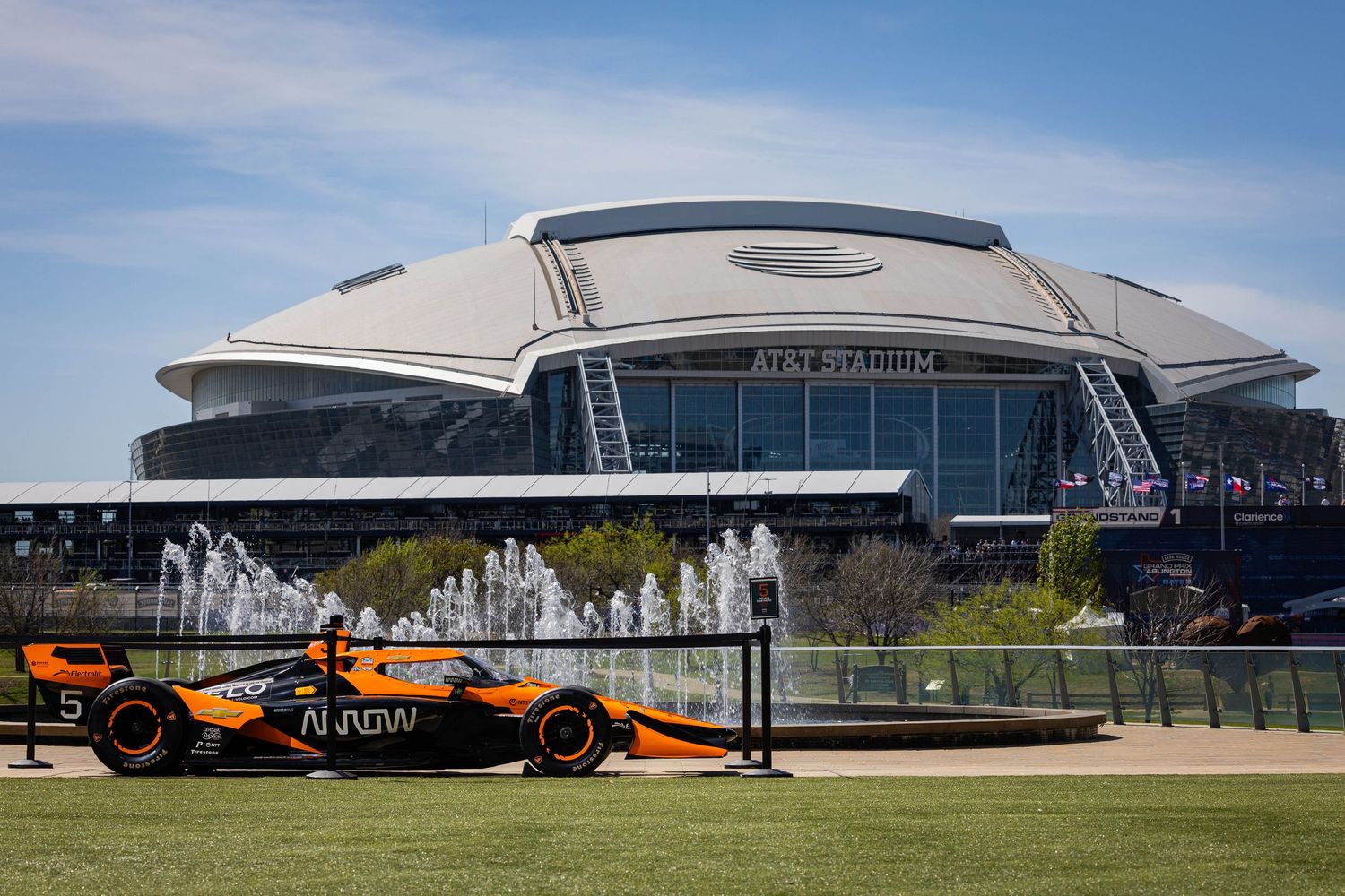 Arrow McLaren orange IndyCar speeds past AT&T Stadium with blue sky and fountains in background.