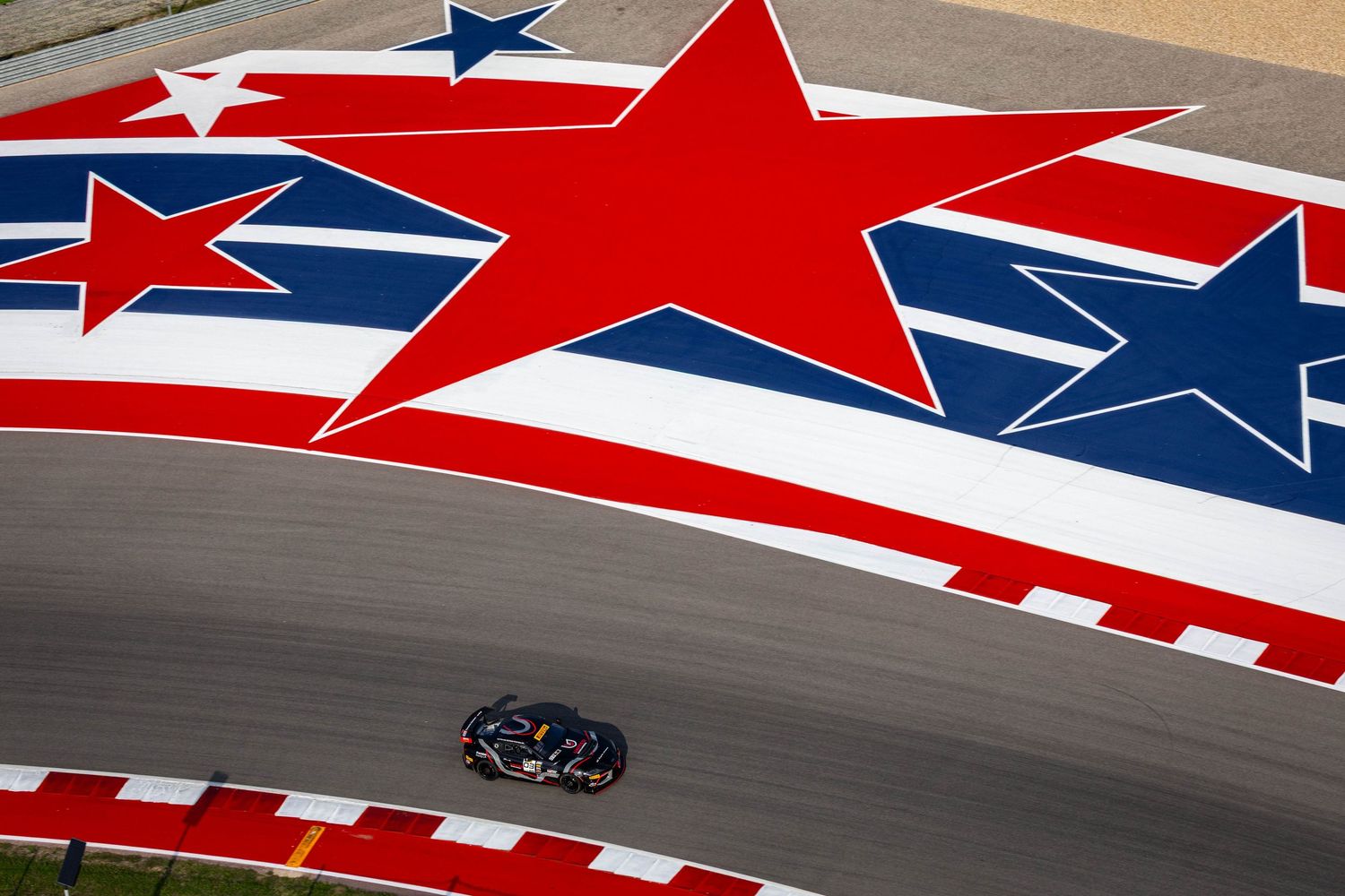 Aerial view of a racing car on track beneath a giant patriotic American stars mural at COTA.