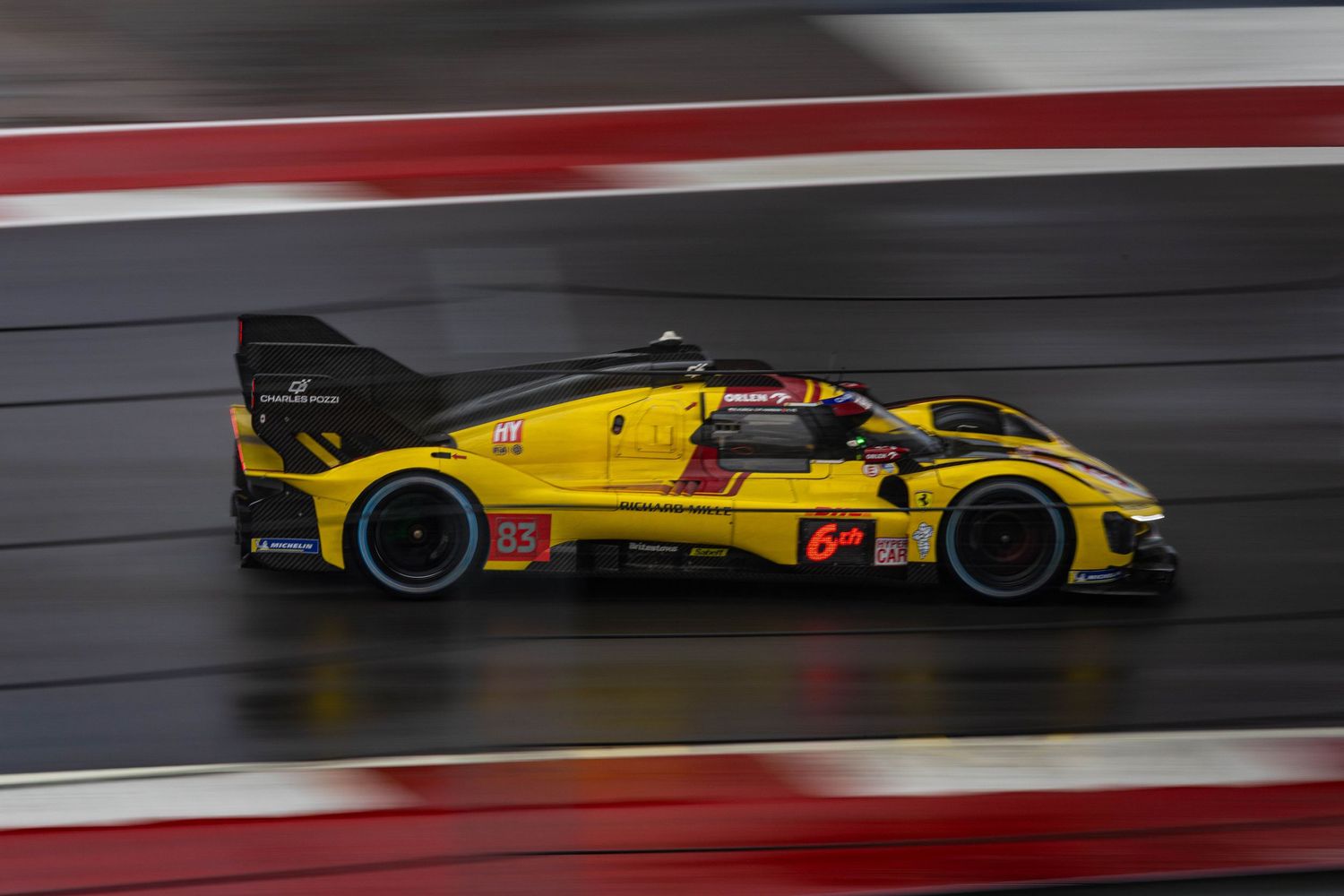 Yellow Corvette GTP prototype endurance race car speeds along wet circuit with motion blur effect.