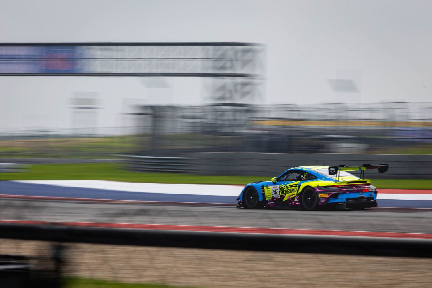 A blue and yellow Aston Martin GT3 race car speeds along a wet professional racing circuit.