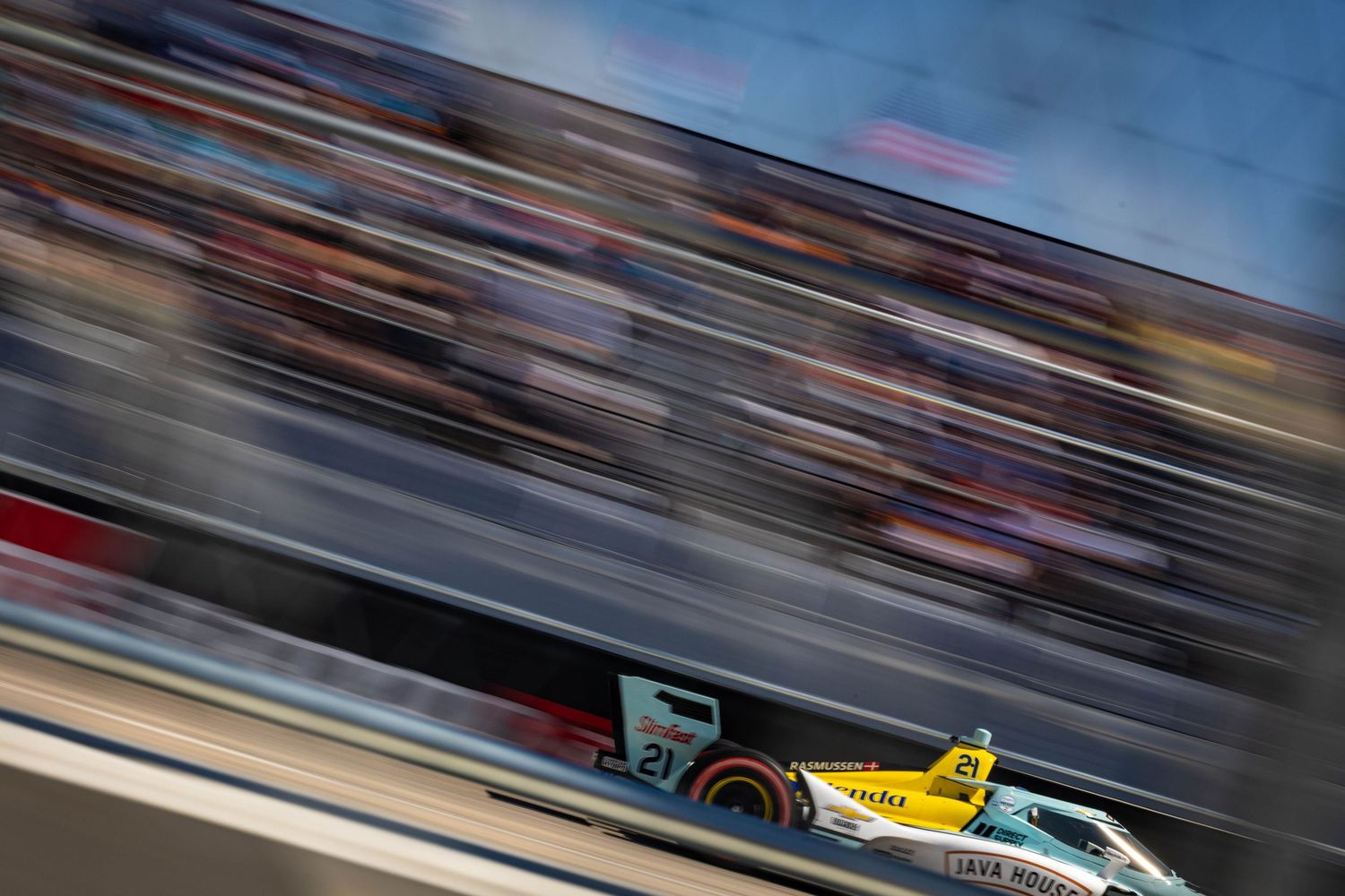 Yellow and green IndyCar speeding past packed grandstands full of racing fans.