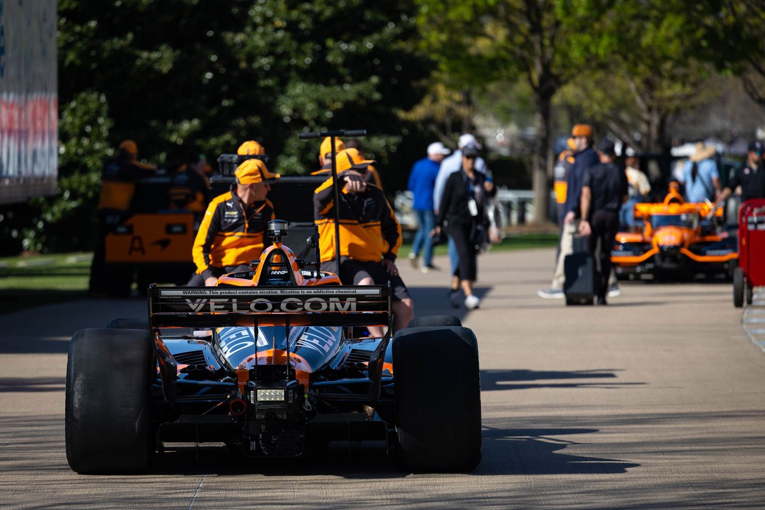 McLaren IndyCar being pushed through paddock by crew members with VELO.com sponsorship visible on the rear wing.