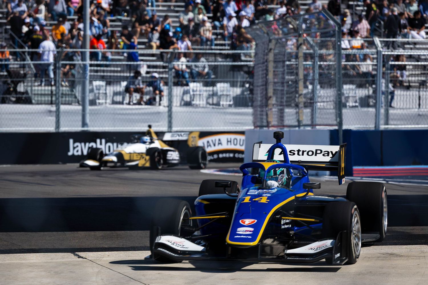 Indy car racing on a street circuit with blue and yellow livery leading a black car past grandstands.