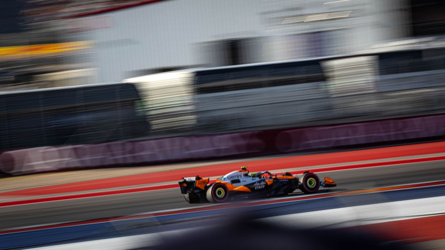 McLaren Formula 1 car in orange livery racing at high speed along a circuit straight with motion blur.