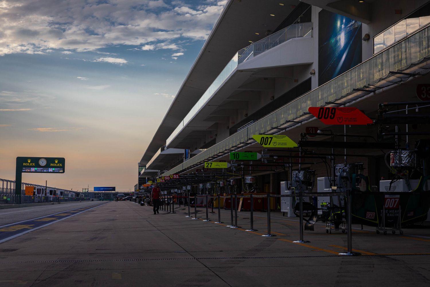 Empty pit lane at a motorsport circuit during golden hour sunset with colorful team umbrellas lining the paddock.