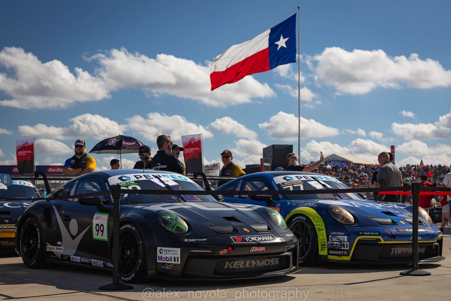 Two Porsche GT3 race cars on display under the Texas state flag at a motorsport event.