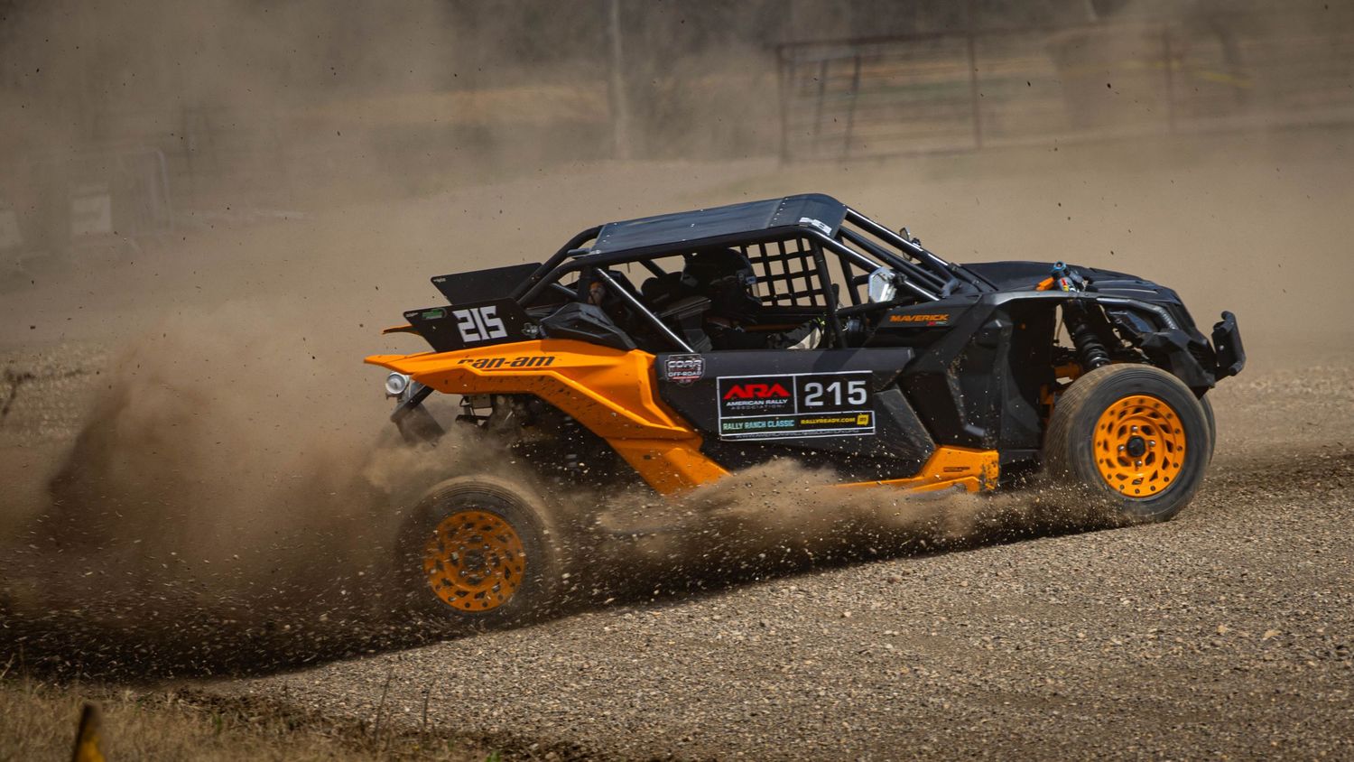 Orange and black off-road race car number 215 kicking up dust on a dirt track during a rally race.