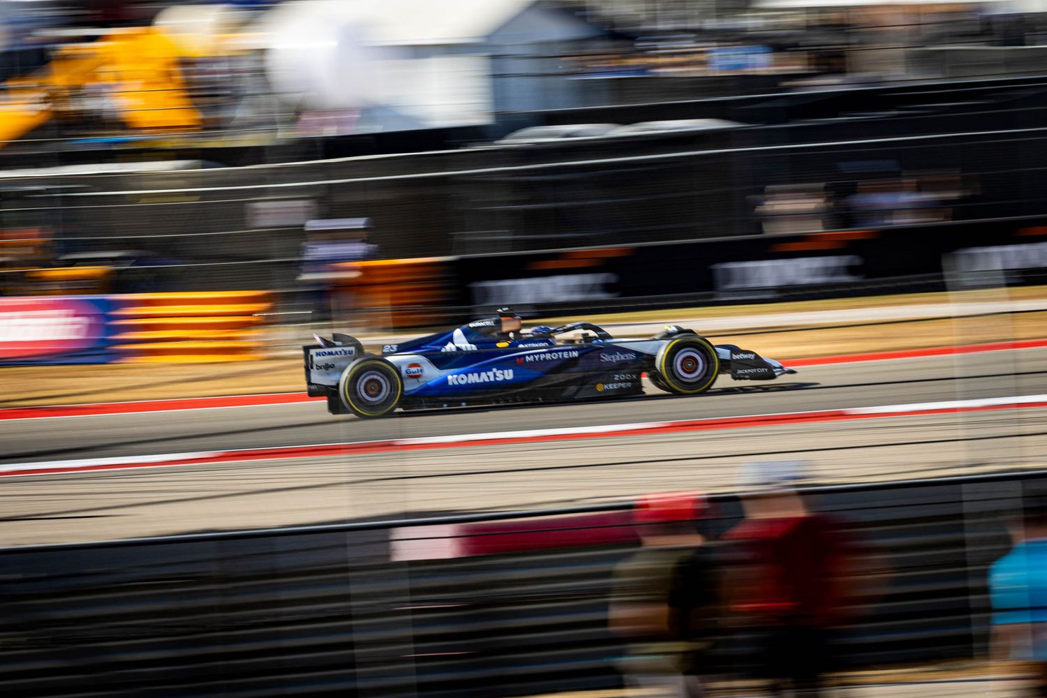 Blue Formula racing car accelerates through pit lane with motion blur at a professional racing circuit.