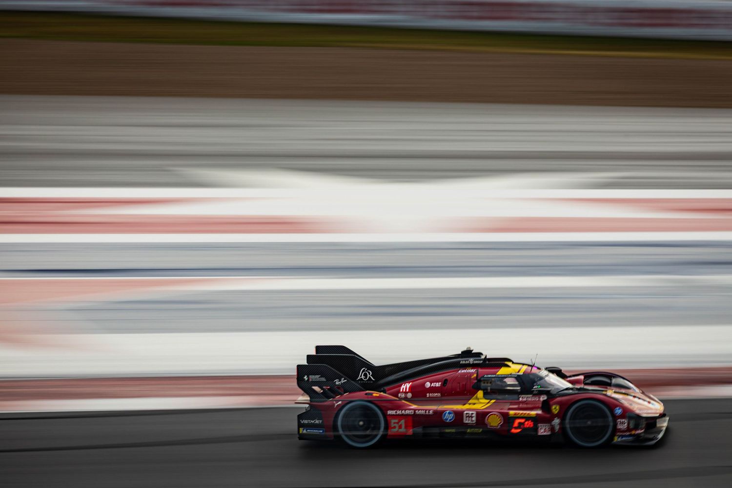 Ferrari LMH prototype race car speeding past blurred grandstands during endurance race.