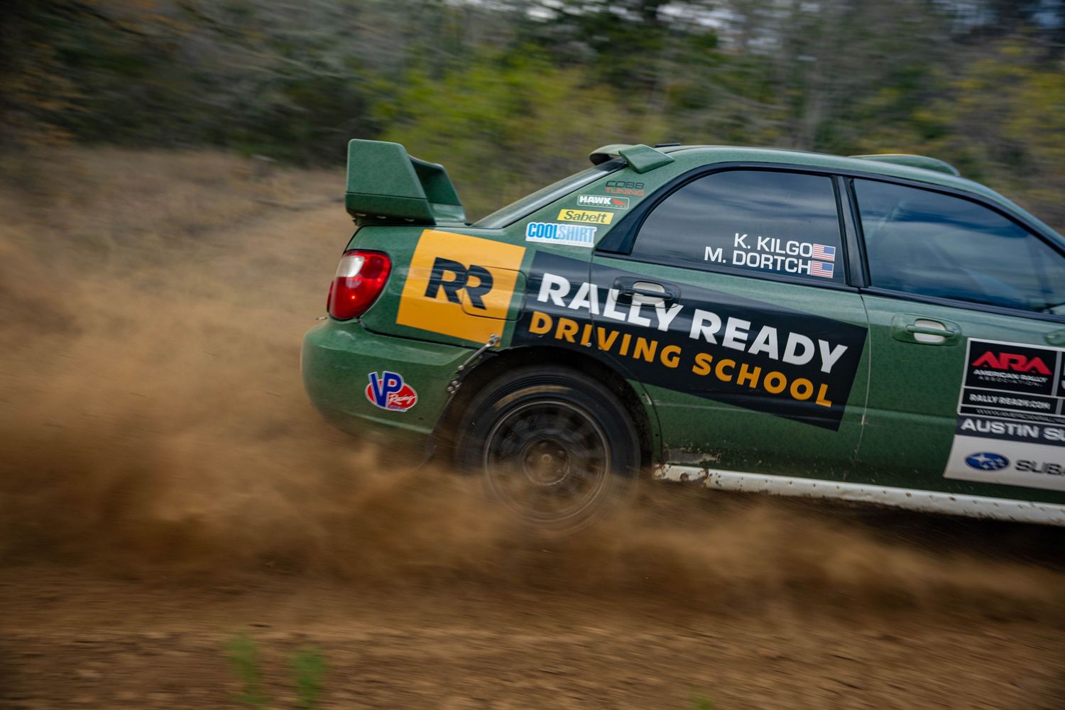 A Rally Ready Driving School Subaru Impreza navigates a dusty gravel rally stage at speed.