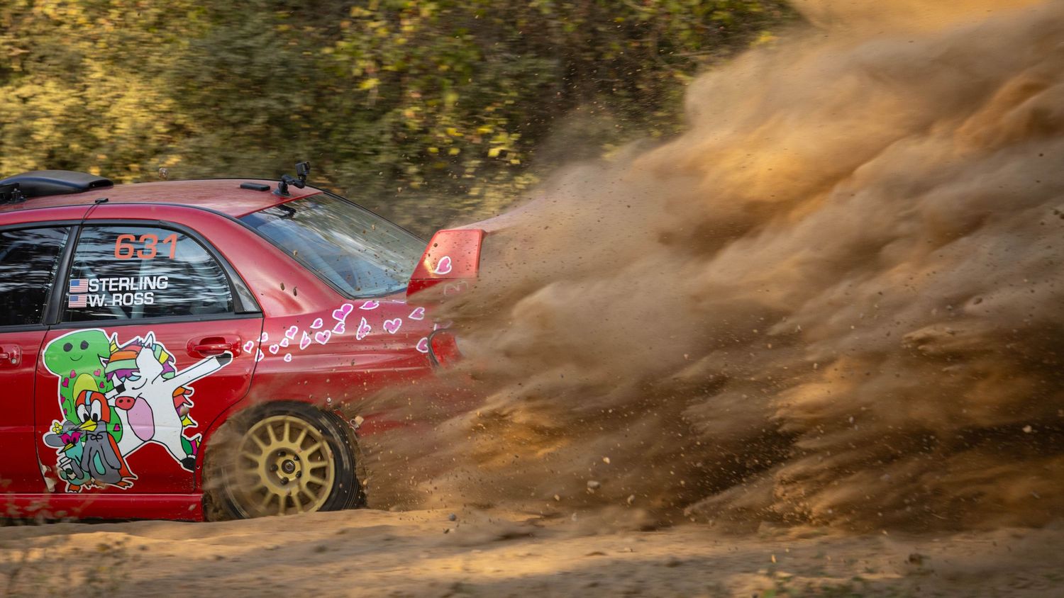 A red rally car with cartoon livery creates a dramatic dust cloud on a dry gravel stage.