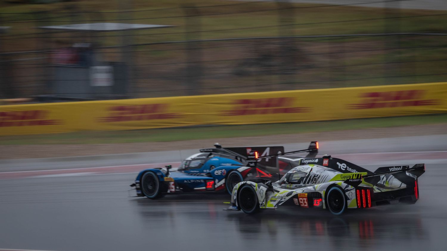 Two prototype endurance racing cars battling side by side on a wet circuit with DHL branding in the background.