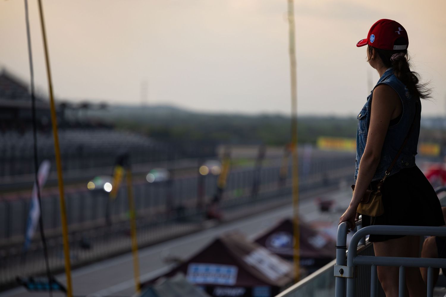 Woman in red cap watches motorsport race from elevated vantage point overlooking circuit at sunset.