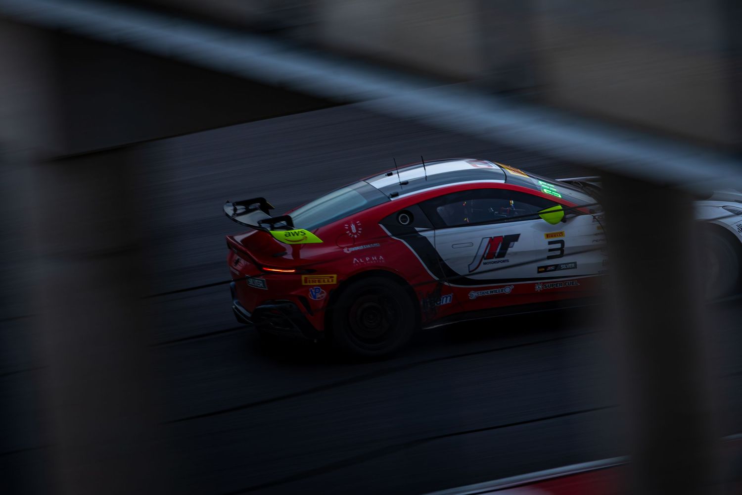 Red Porsche 911 GT3 race car captured through fence in motion during a competitive motorsport event.