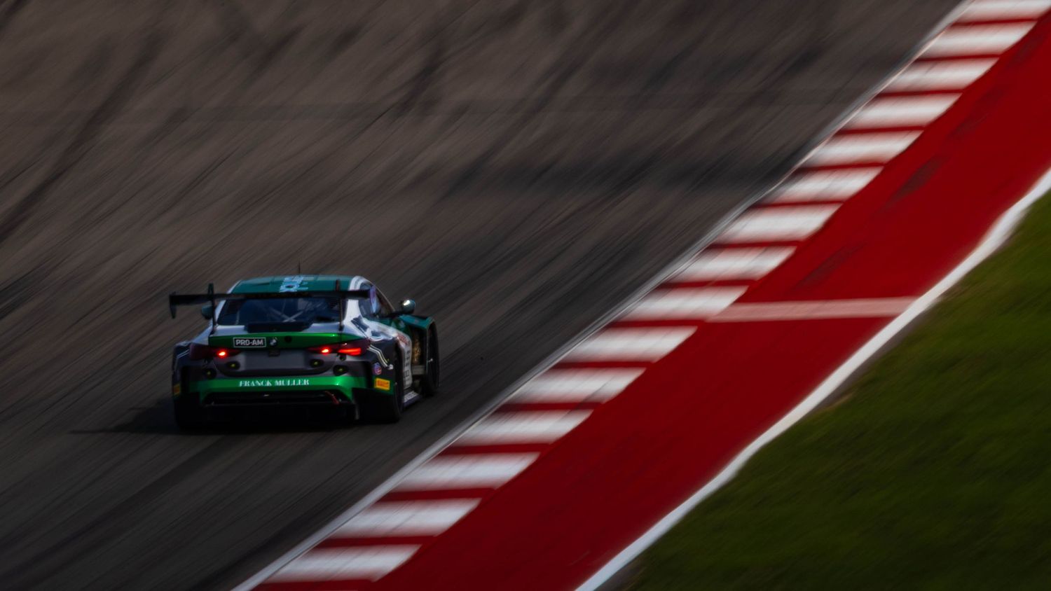 A dark green GT racing car speeds through a corner at dusk on a professional race circuit.