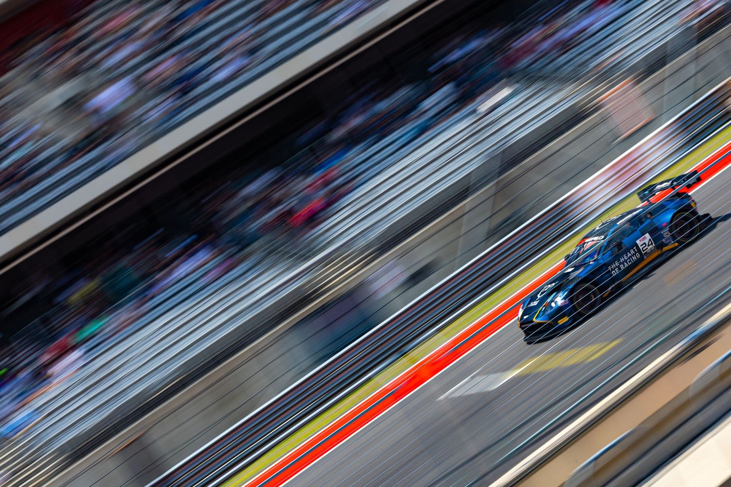 Blue GT racing car speeds past packed grandstands at a motorsport circuit, captured with motion blur effect.