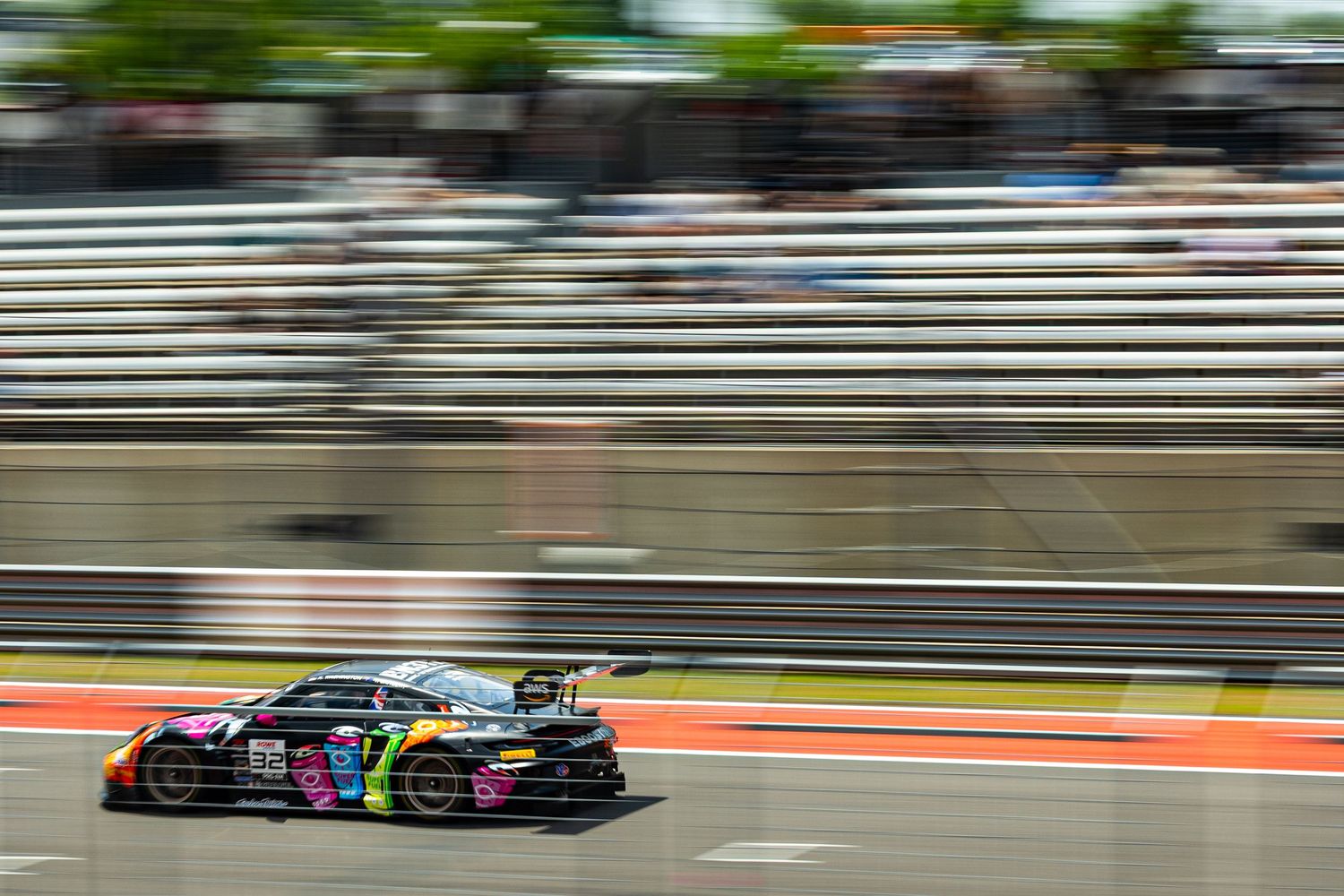 Rainbow-liveried GT3 race car speeding past grandstands on a sunny race circuit.