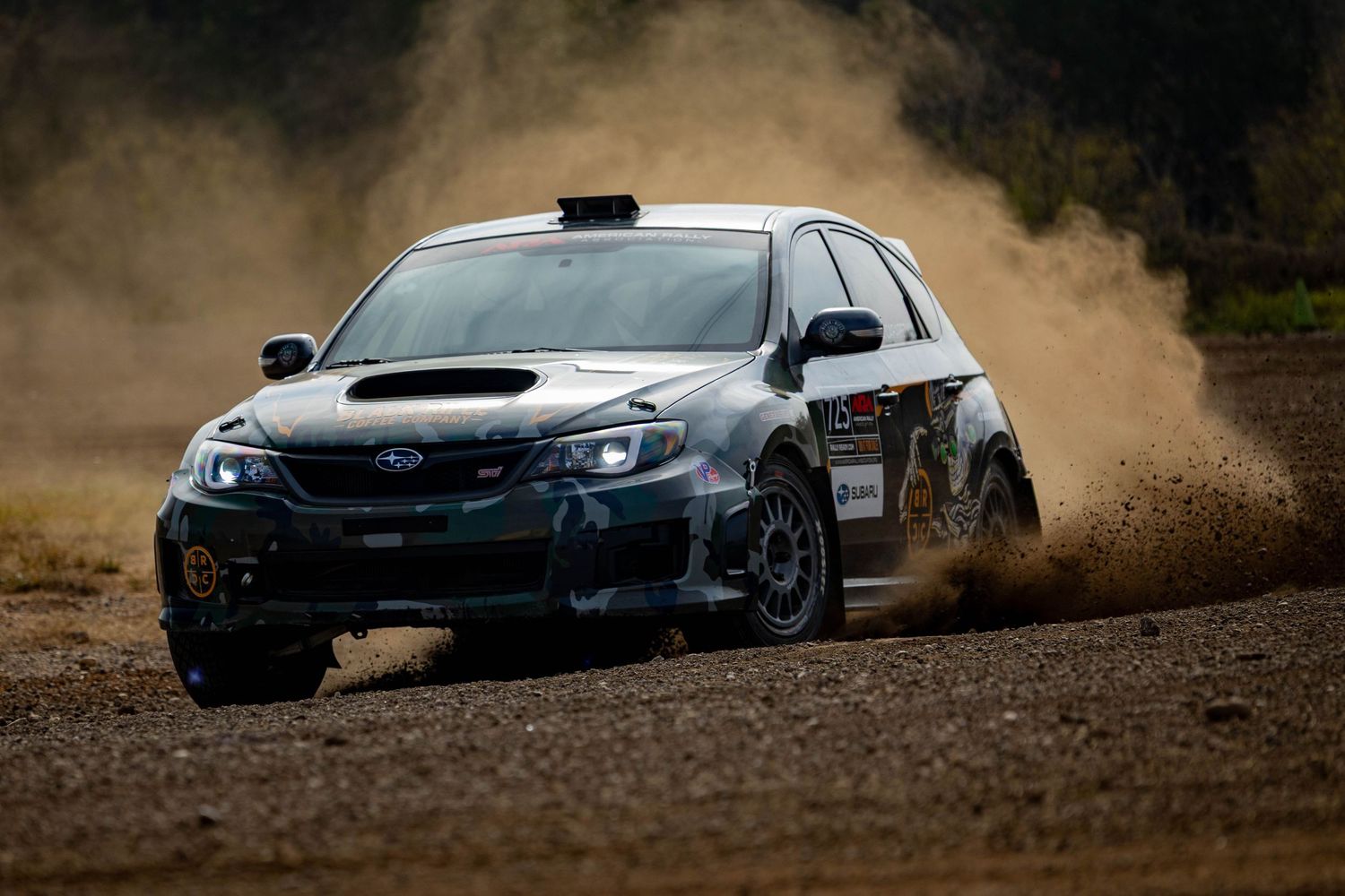 A Subaru WRX STI rally car kicks up dust while cornering on a gravel rally stage.
