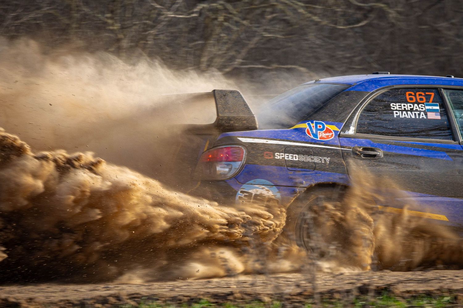 Blue rally car number 667 launching massive dirt spray on a muddy off-road rally stage.