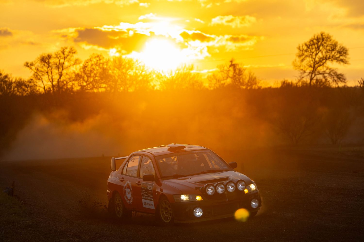 Rally car racing on a dirt road at sunset with golden light and dust clouds.