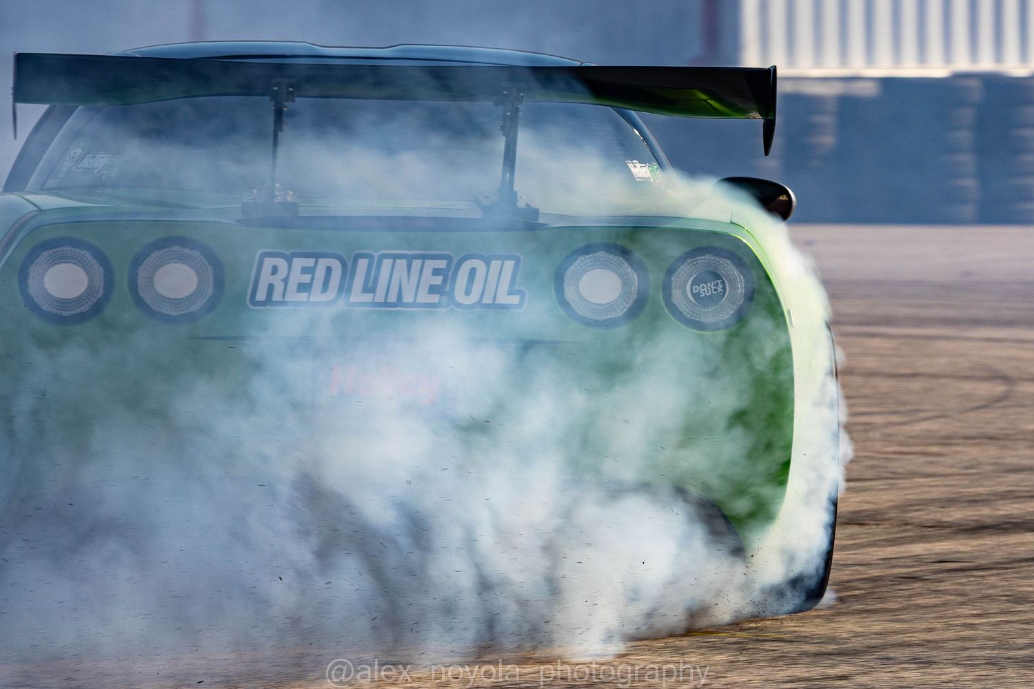 A green racing car with Red Line Oil livery performing a burnout, surrounded by thick white tire smoke on a race track.