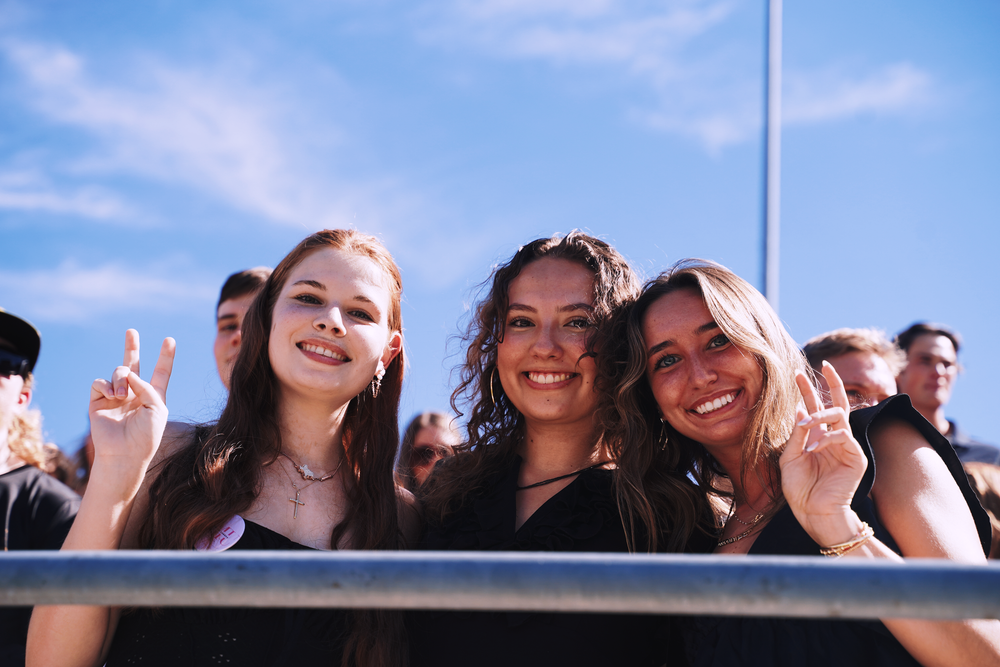 Three young women smile and make peace signs at an outdoor music festival under a blue sky.