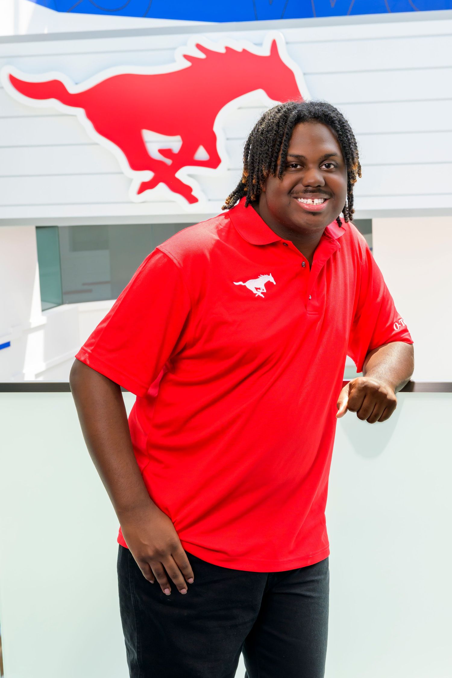 A smiling student in a red SMU Mustangs polo shirt poses in front of the school logo.