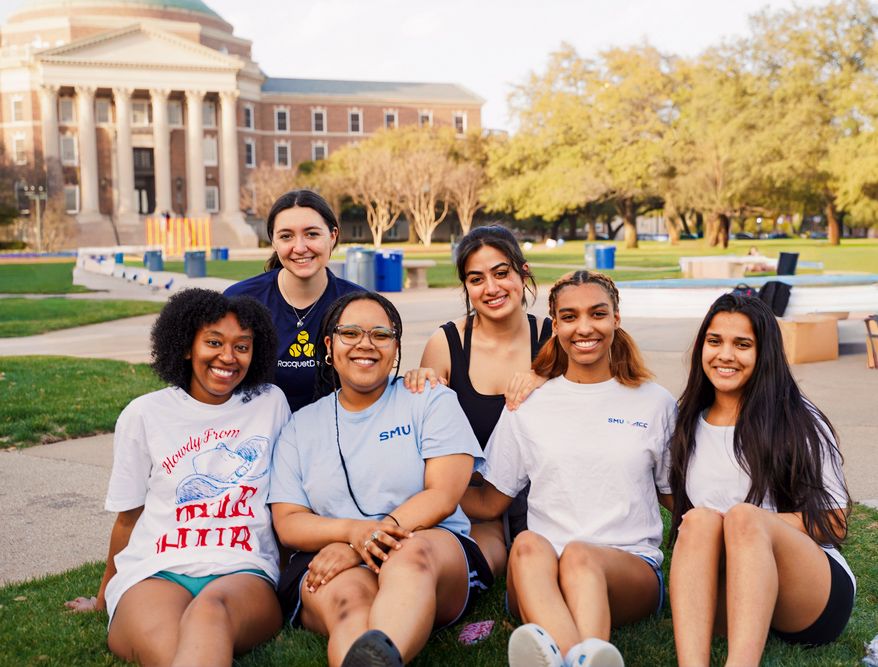 Six diverse college women pose together smiling on a university campus lawn.