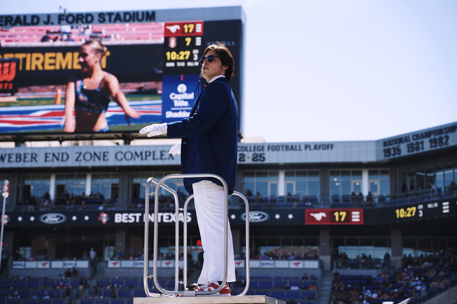 A man in a blue blazer speaks at a podium during a college football playoff game at Gerald J. Ford Stadium.