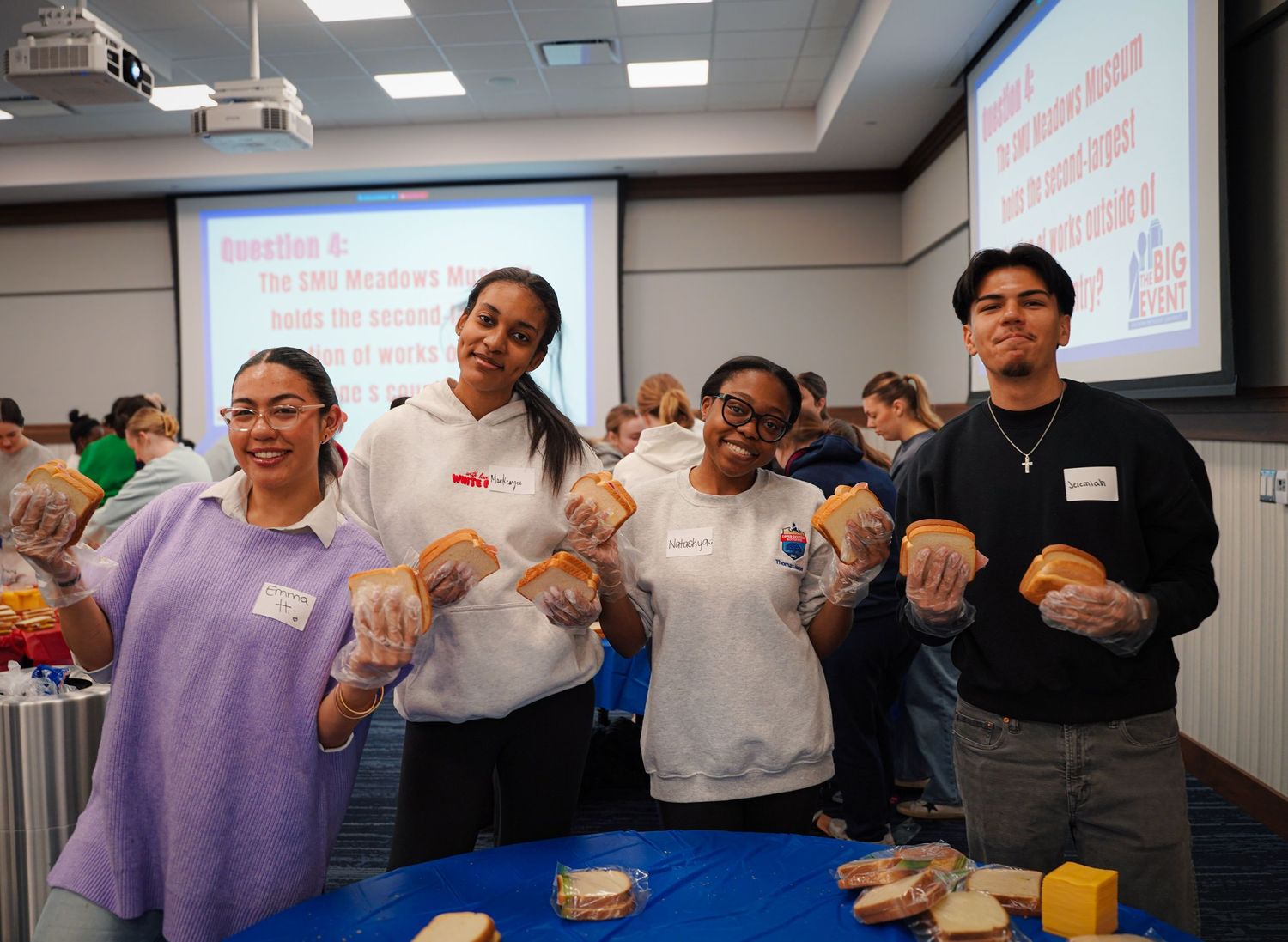 Four students hold bread rolls while participating in a food assembly volunteer activity indoors.