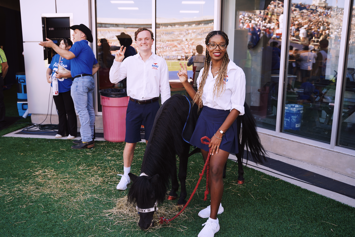 A woman with braids holds a black dog on a red leash at an outdoor SMU campus event with a man in a white shirt.