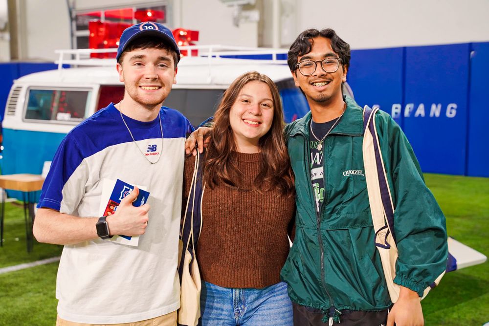 Three college students smile together indoors at an SMU campus event near a vintage blue van.