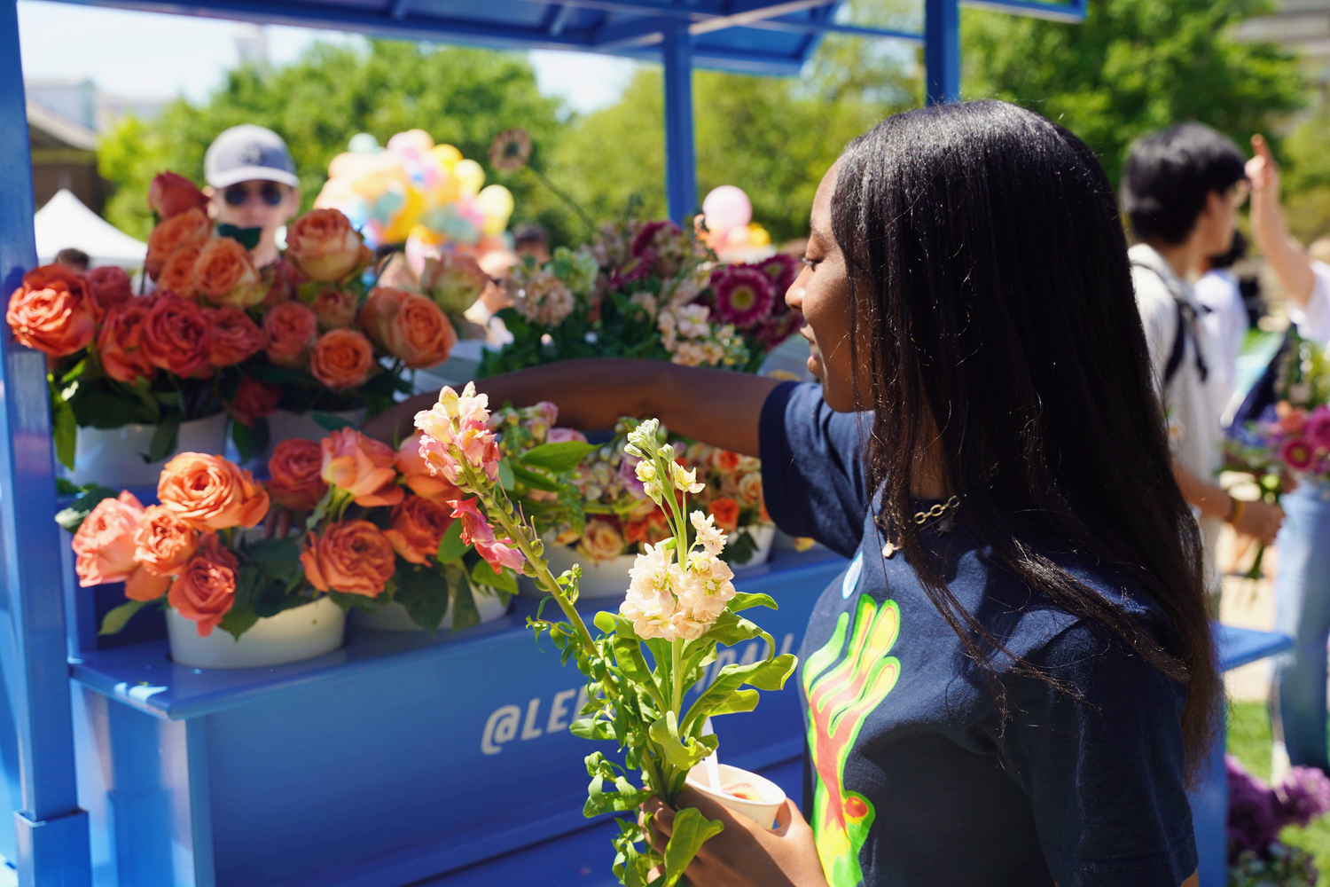 A student browses colorful orange and pink roses at an outdoor flower stand on a sunny campus day.