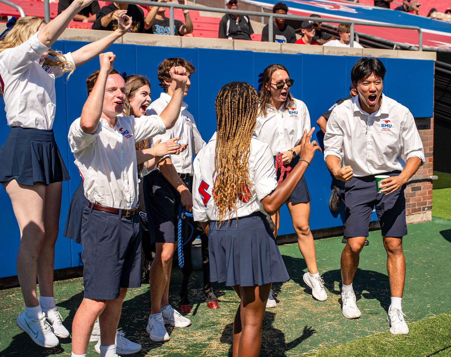 Excited students in white uniforms celebrate outdoors on a sunny day at a campus sporting event.