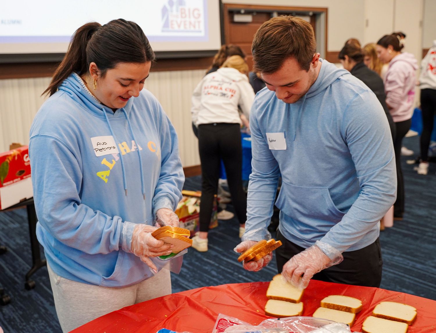 Two students in matching blue hoodies assembling sandwiches at a table during a campus volunteer service event.