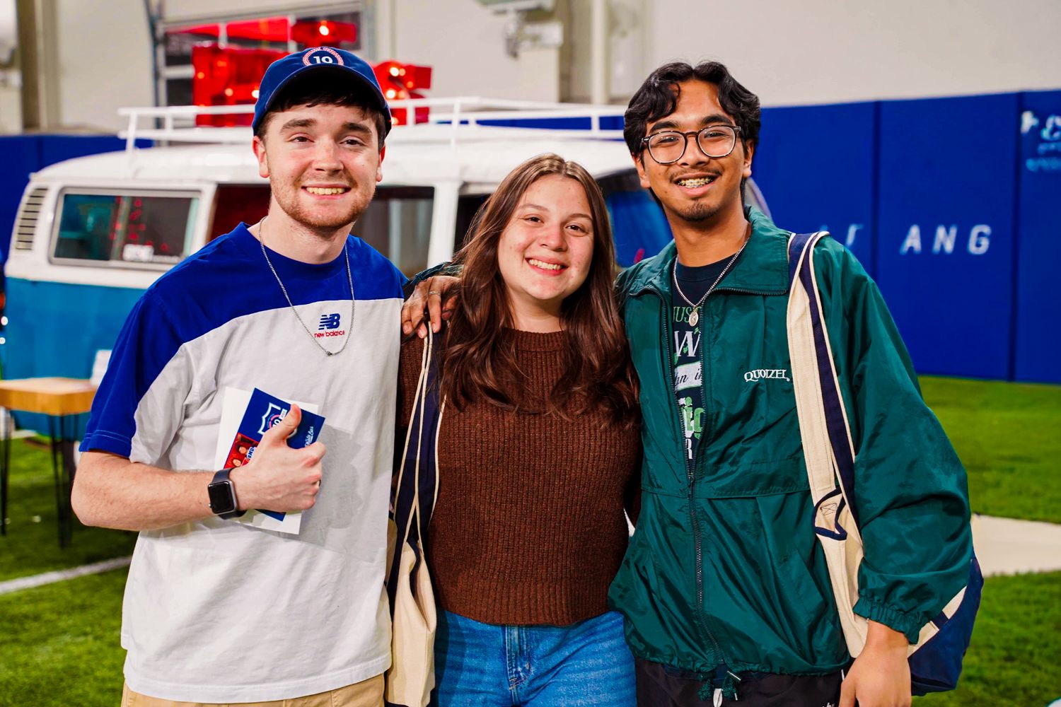 Three smiling college students pose together indoors near a blue vintage van at a campus event.
