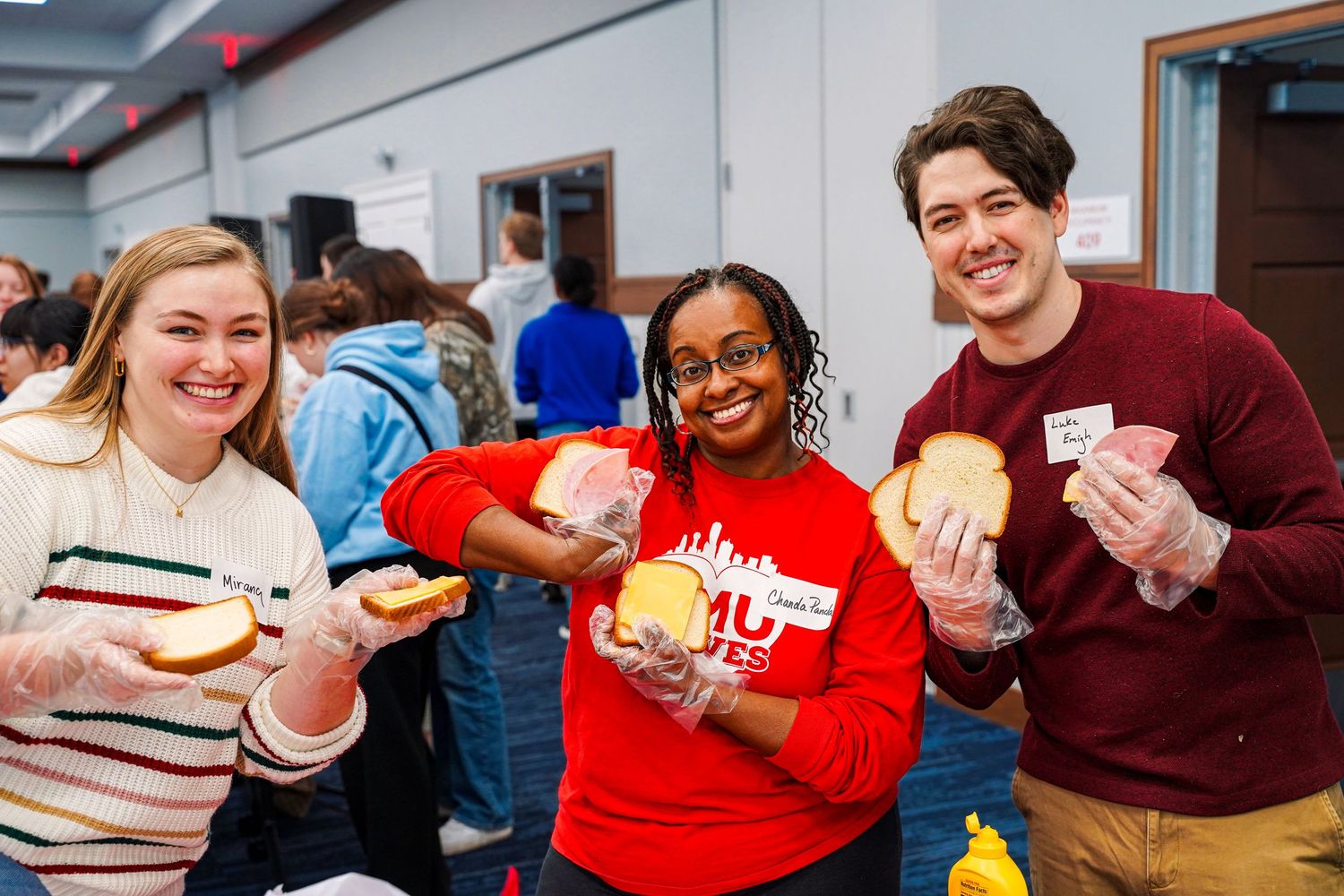 Three college students smiling and holding sandwiches during a university community service volunteer event.