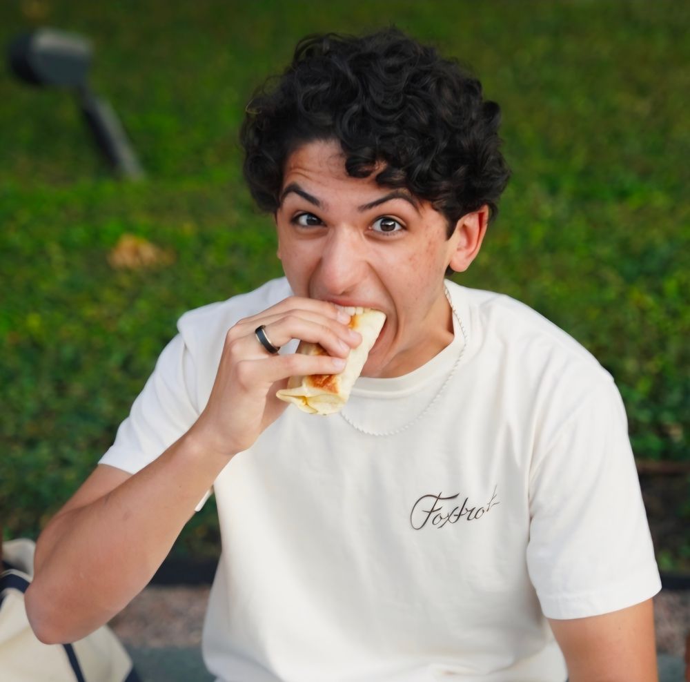 Young man with curly hair eating a sandwich outdoors on green grass.