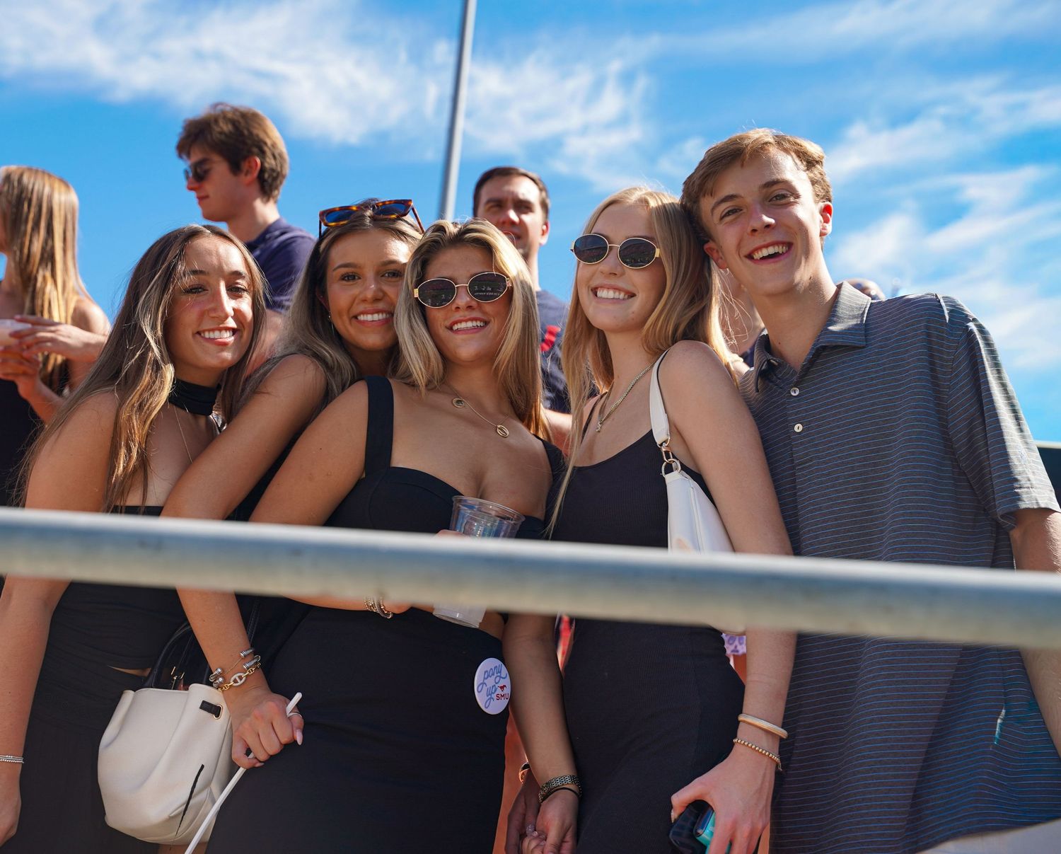 Group of smiling young adults posing together outdoors at a sunny event under a blue sky.