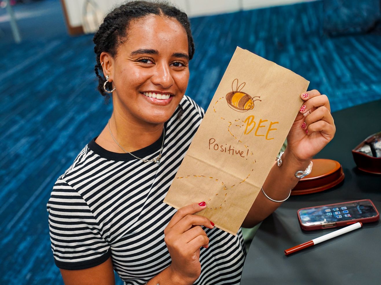 Smiling young woman holding a brown paper bag decorated with Bee Positive message and bee drawing.
