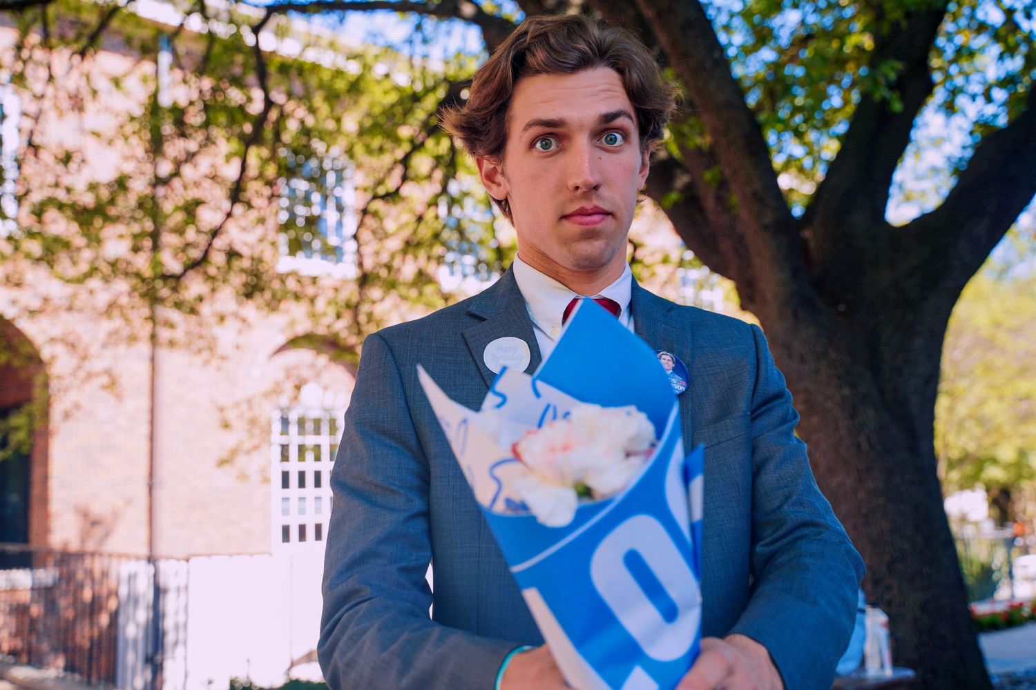 Young man in a grey suit holds a blue cotton candy bag outdoors near a tree on campus.