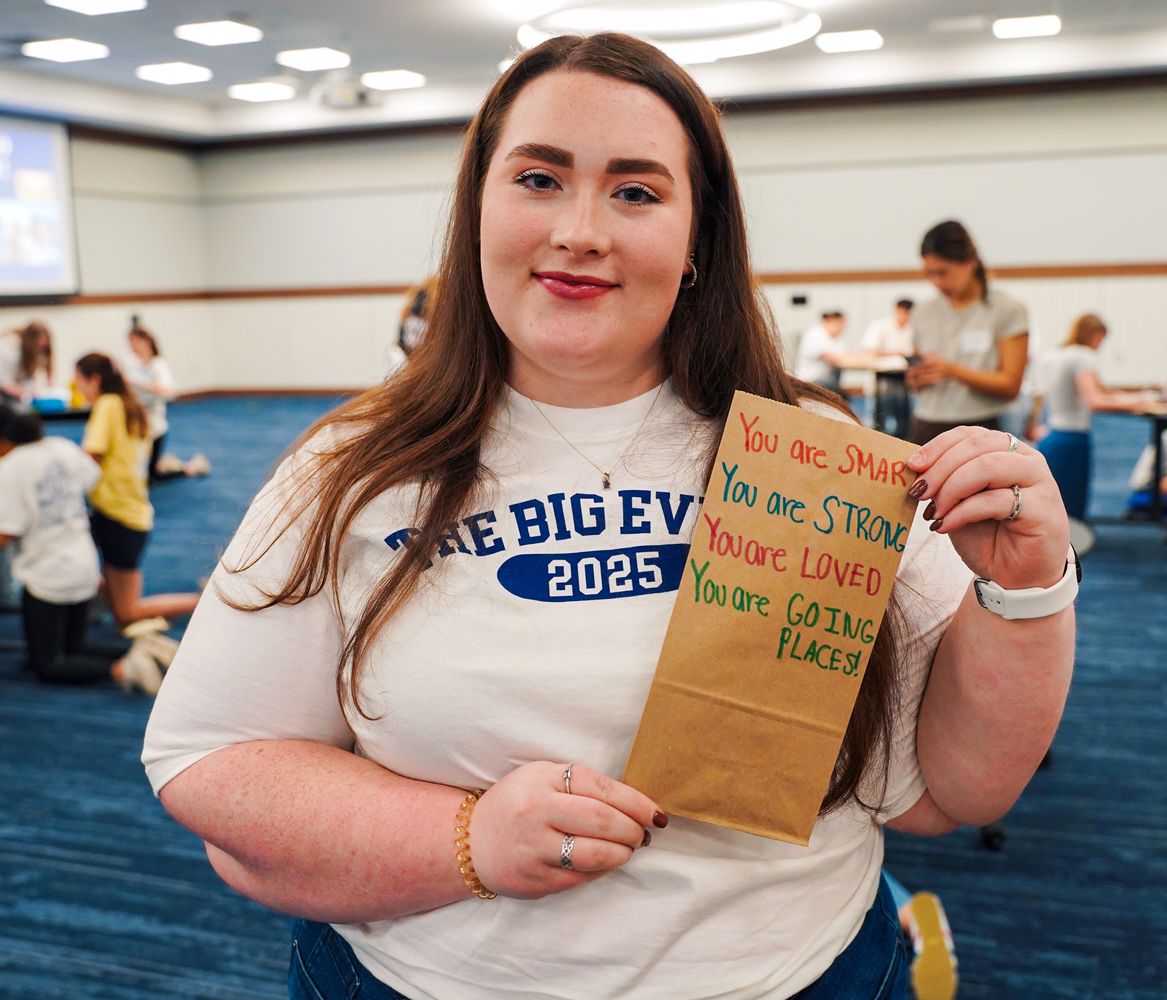 Smiling college student holds a decorated paper bag with encouraging words at a campus event.