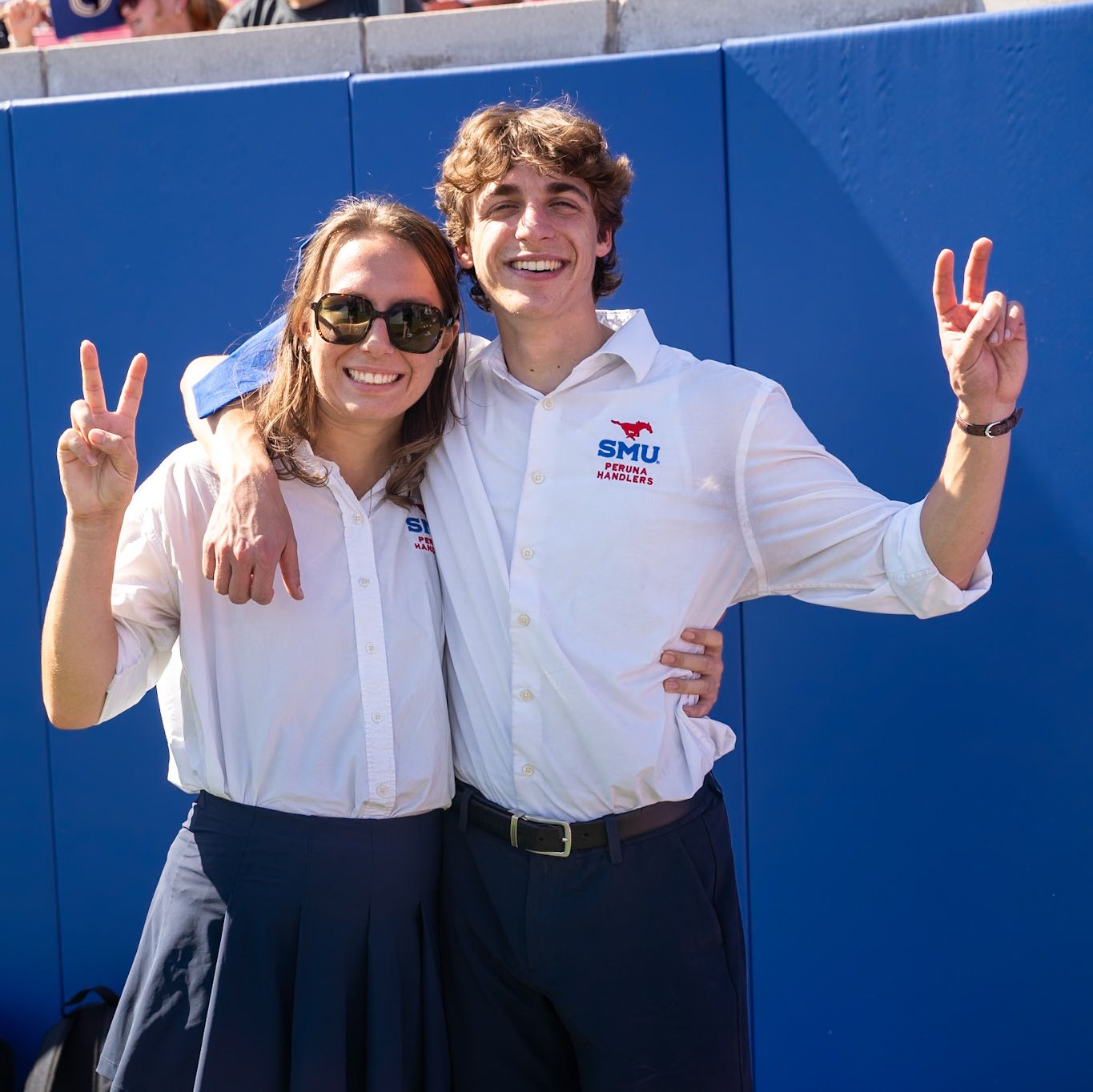 Two SMU student ambassadors in white uniforms flash peace signs at a sunny outdoor stadium event.