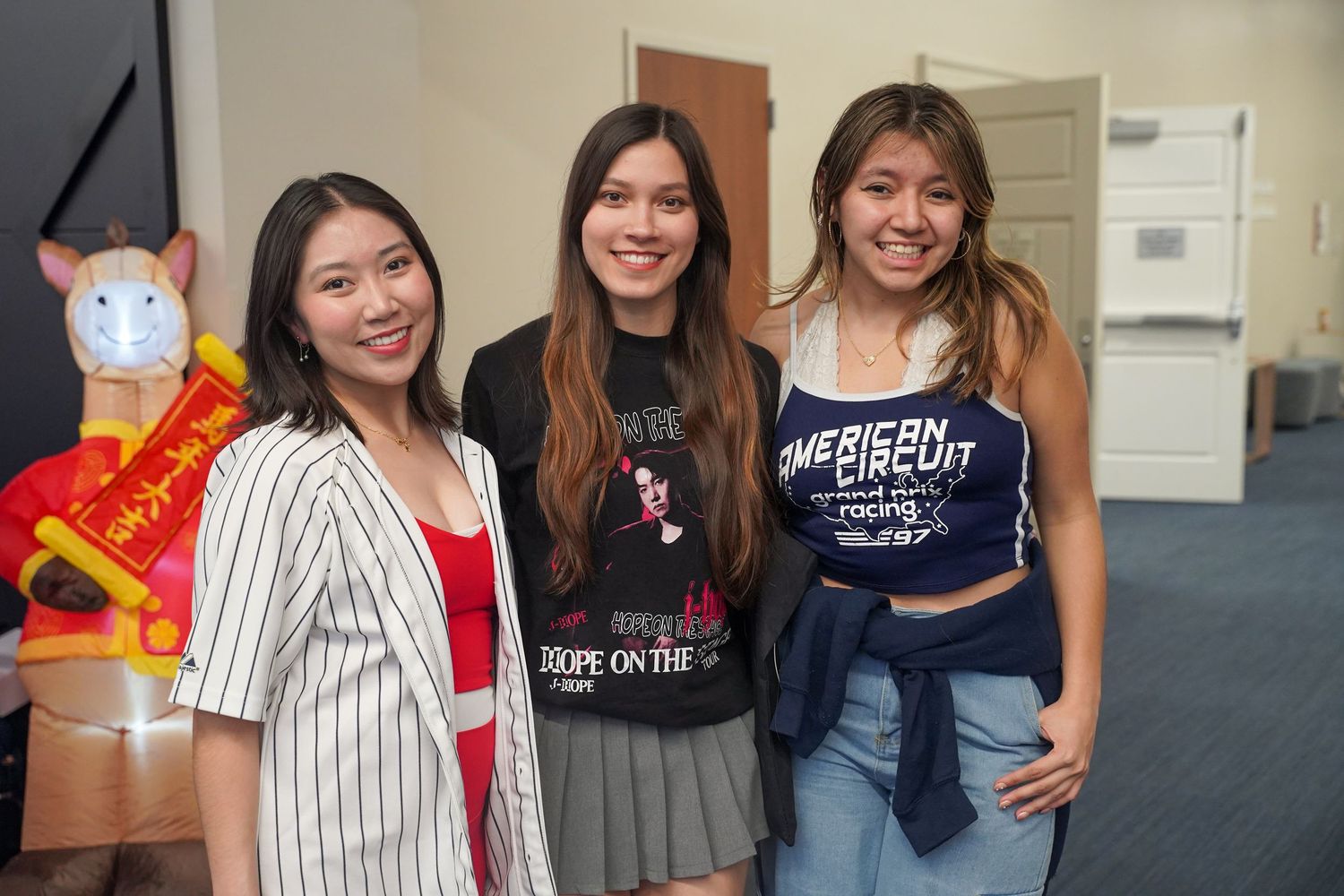 Three young women smile together at an indoor campus cultural event with festive decorations nearby.