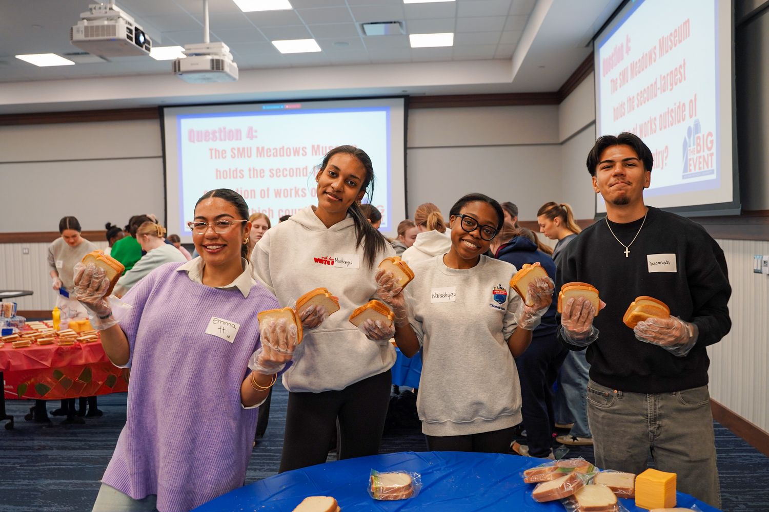 Four diverse college students smiling while holding sandwiches during a community service sandwich-making event.