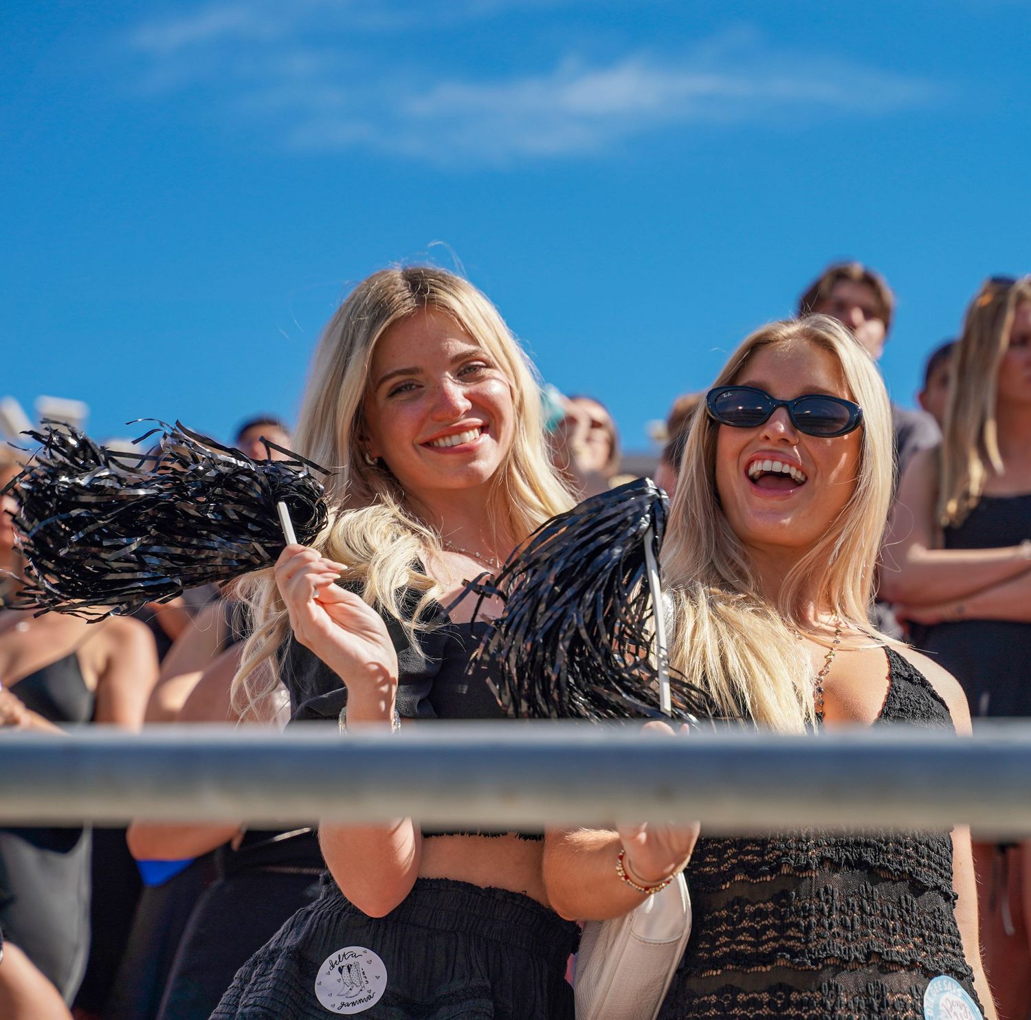 Two smiling blonde women holding black pom-poms at an outdoor stadium event under a bright blue sky.