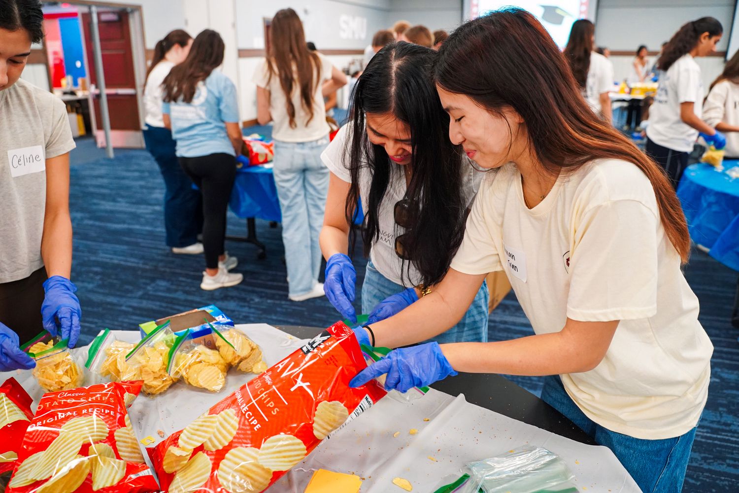 Two female students wearing blue gloves pack food bags at a volunteer service event.