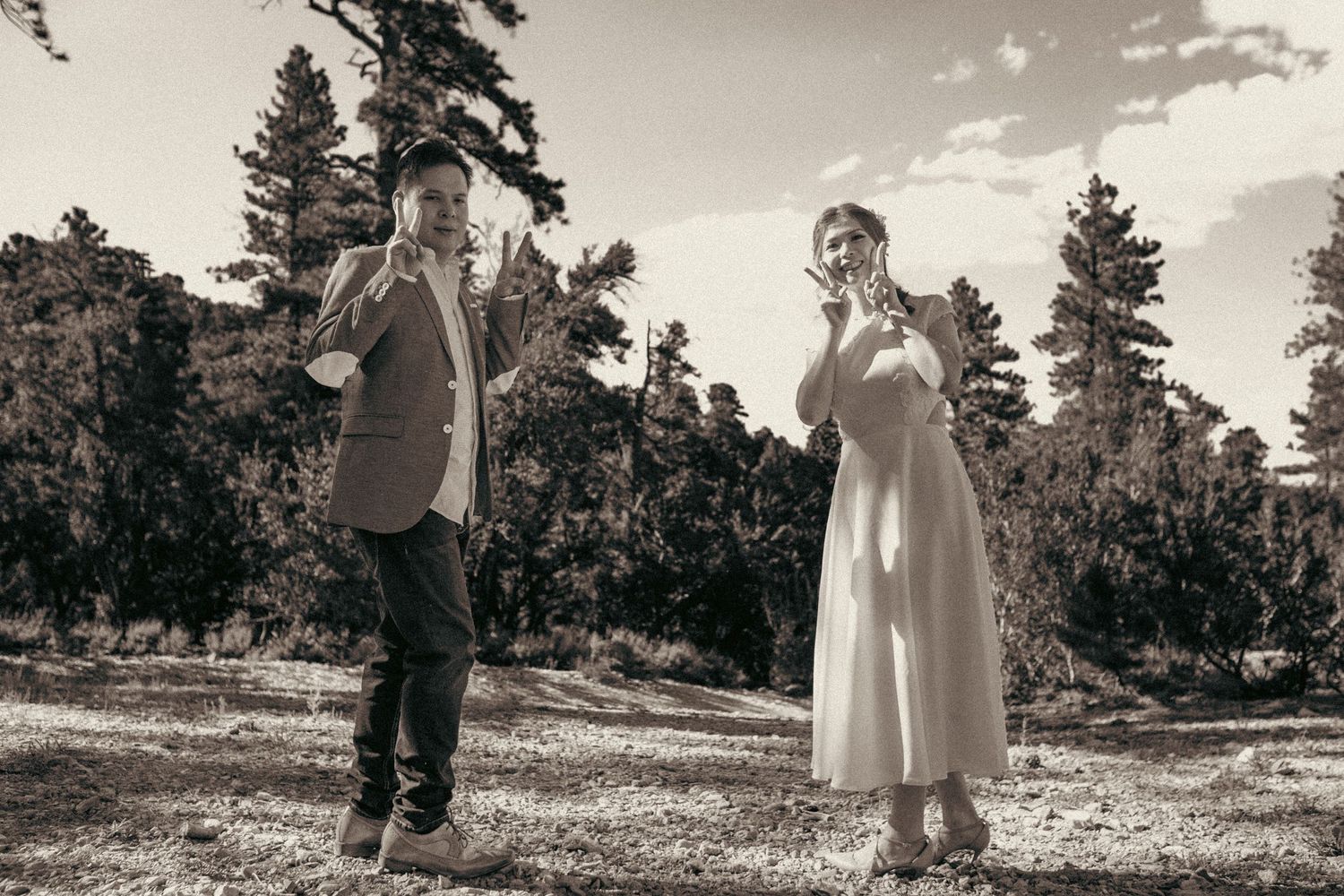 Couple in sepia-toned outdoor photo, man in blazer and woman in white dress among pine trees.